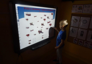 Chłopiec stoi pod tablicą interaktywną przelicza ilość wybranego elementu na obrazku.